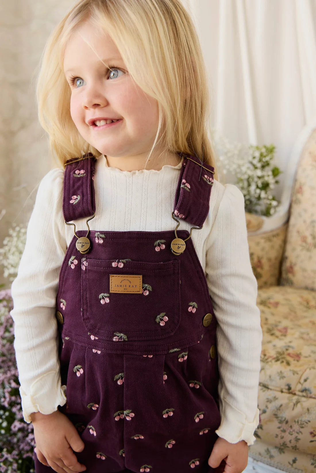 Young girl wearing a purple floral dress over a white shirt, standing in a softly lit room.