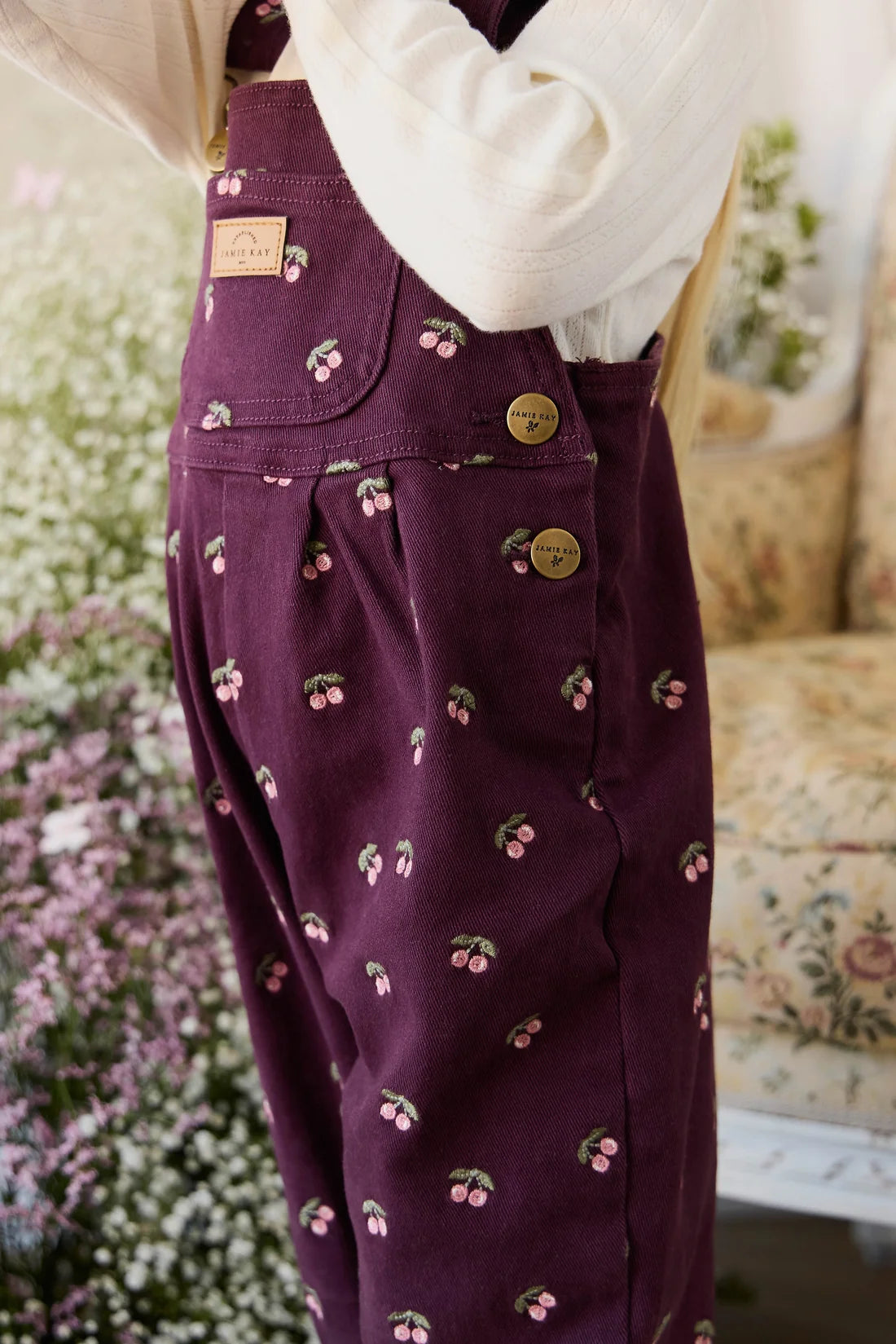 Purple overalls with cherry pattern worn by a person, blurred floral background