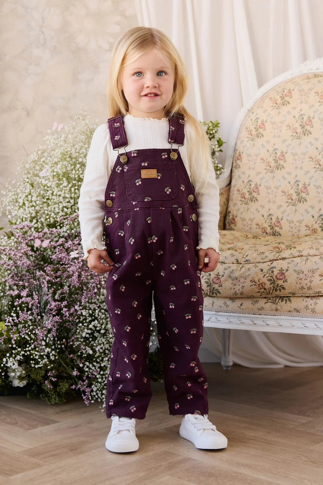 Young girl wearing a white shirt and purple patterned overalls standing in a room with floral decor.