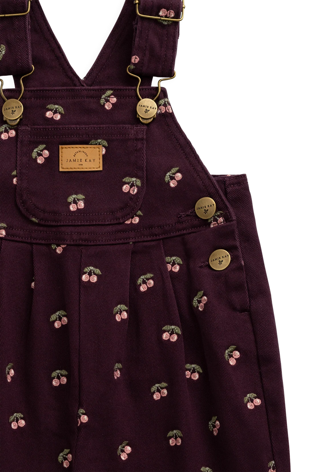 Close-up of a dark purple bib overalls featuring a cherry print pattern, gold-tone metal buttons, and a tan leather brand patch on the chest pocket.
