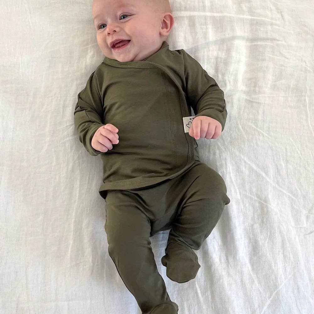 A baby lying on a white textured blanket, wearing an olive green long-sleeve footed sleeper with a front zipper.