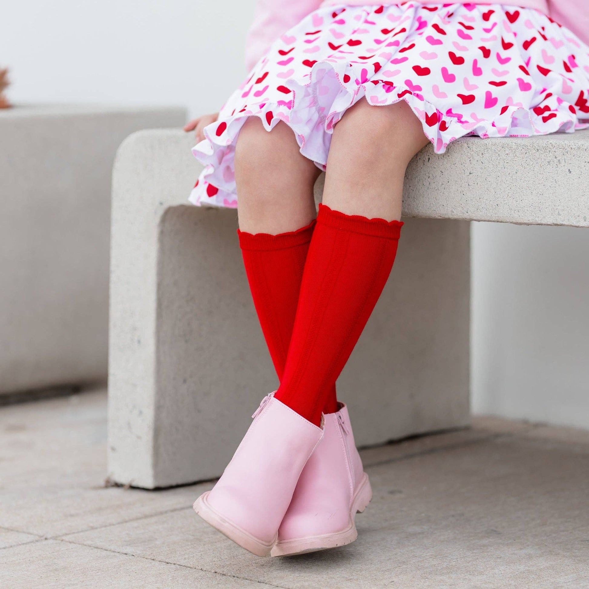 Child wearing red knee-high socks and pink boots sitting on a concrete bench.