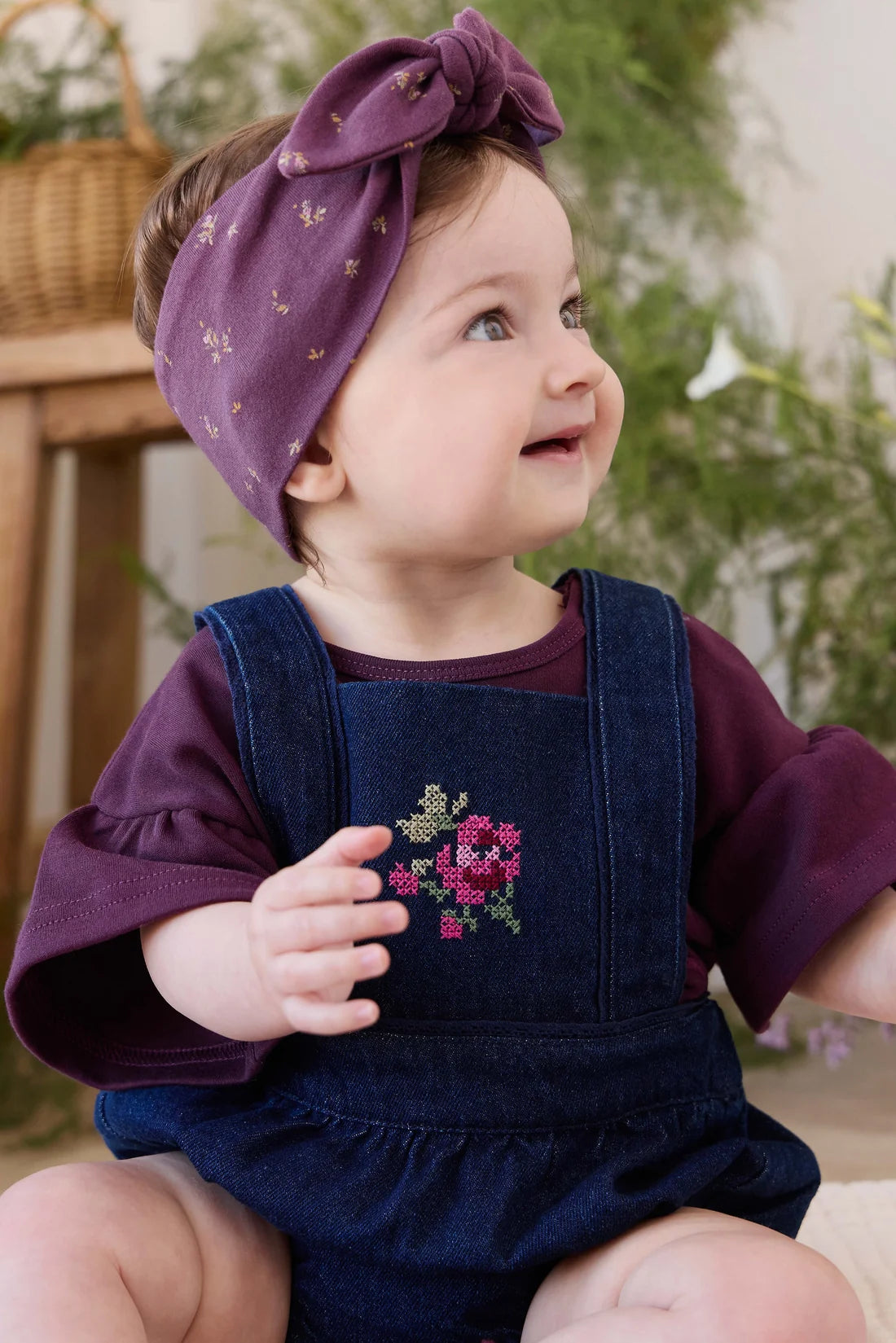 A baby girl wearing a dark purple long-sleeve top, dark blue denim overall shorts with a pink and green embroidered floral design on the chest, and a matching purple fabric headband with small gold stars.