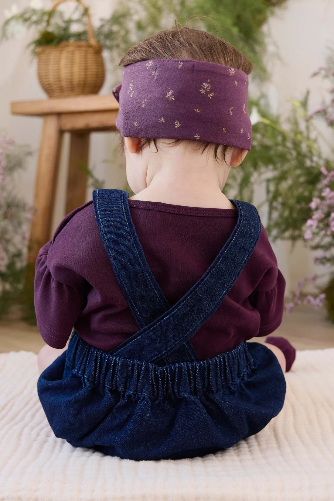 A baby seen from behind wearing a purple long-sleeve top, dark blue denim-style overalls with crossed straps, and a matching purple headband with small gold floral details, seated on a cream textured blanket in a softly styled indoor setting.