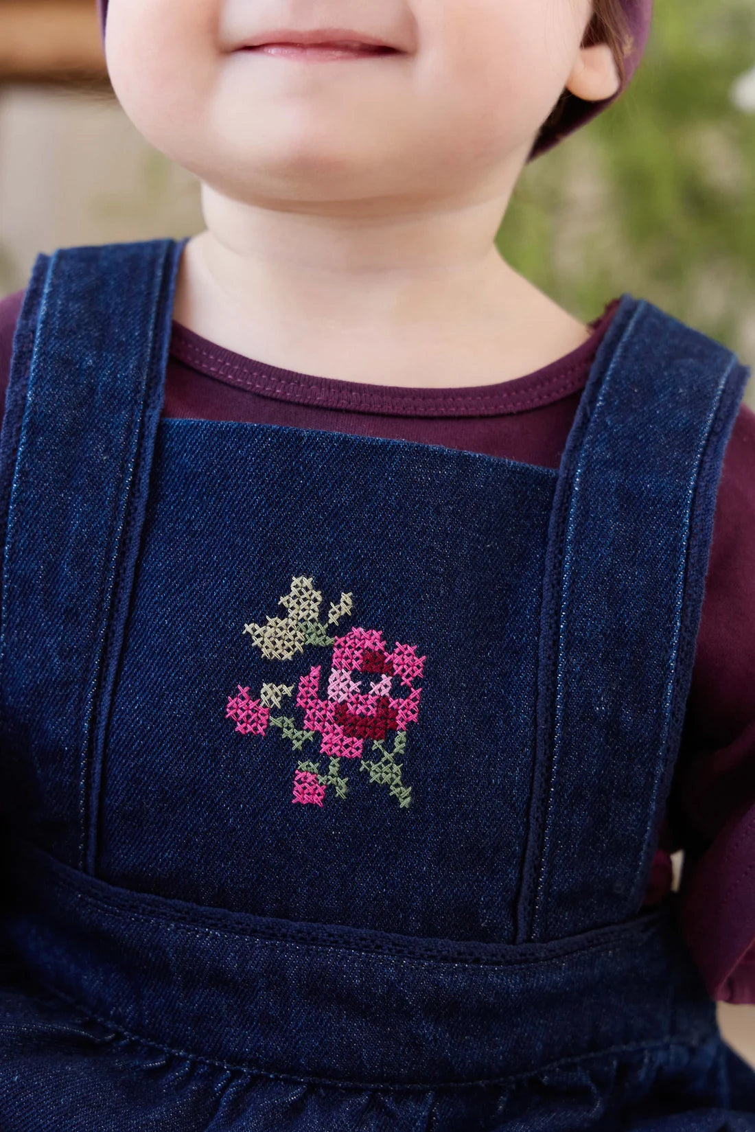 Close-up of a child wearing dark blue denim overalls with a pink and green embroidered rose on the chest over a burgundy shirt.