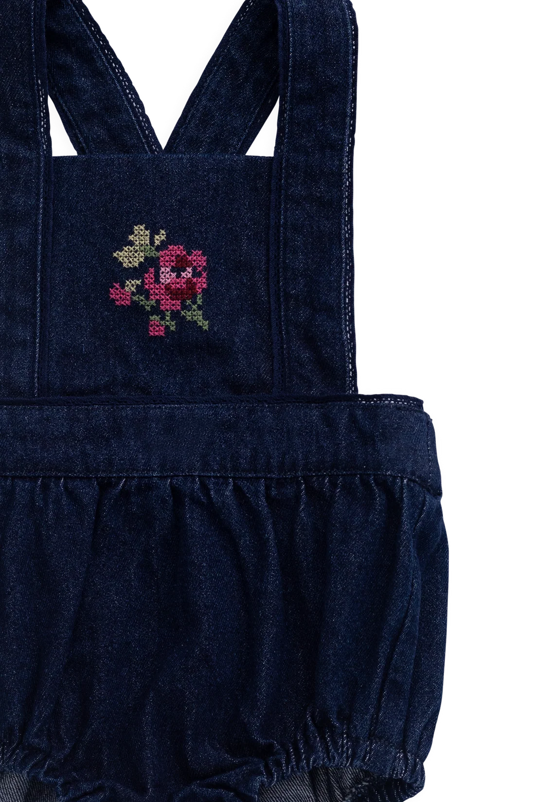 A close-up of a dark blue denim baby romper with a ruffled hem and shoulder straps, featuring a pink and green embroidered rose on the chest pocket.