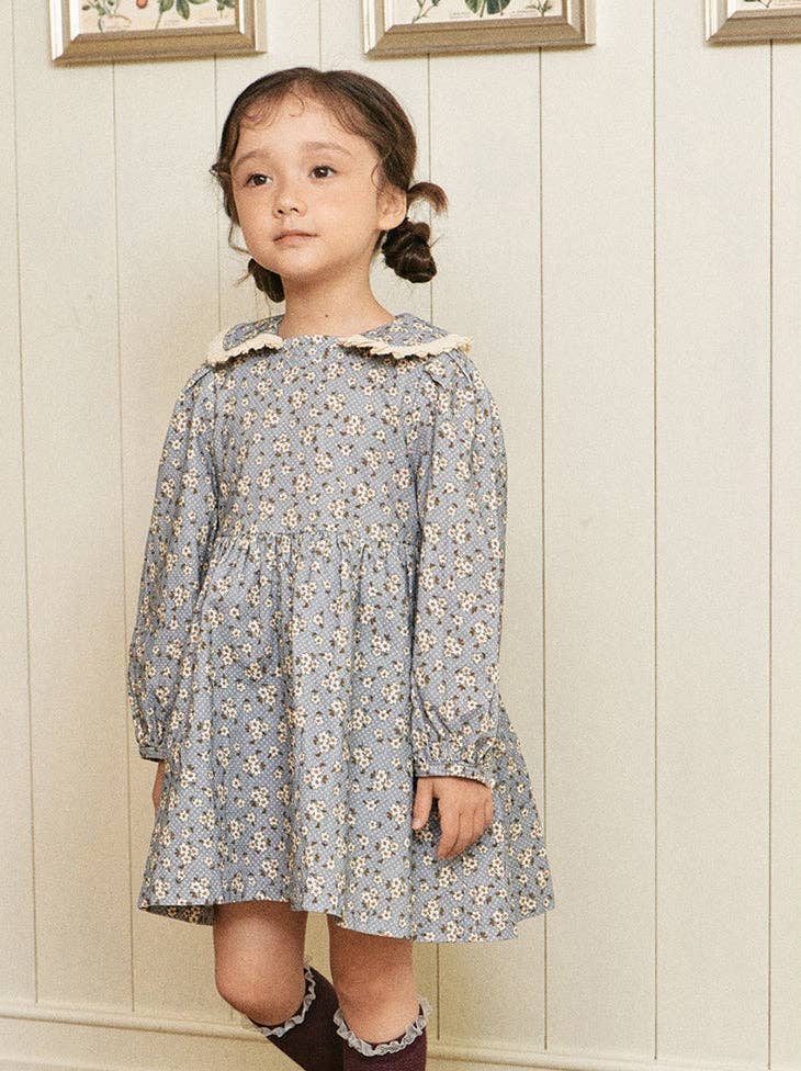 Young girl wearing a floral dress against a light wooden paneled wall.