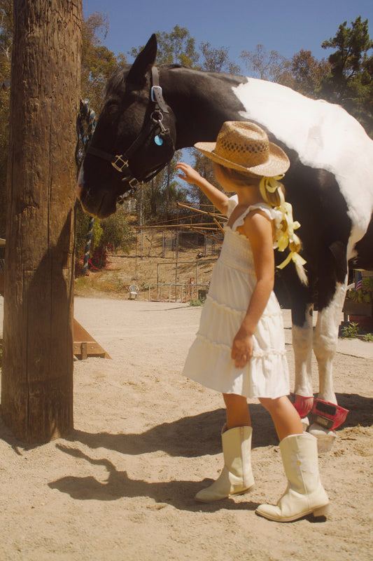 Young girl in a white dress and cowboy hat standing next to a black and white horse.