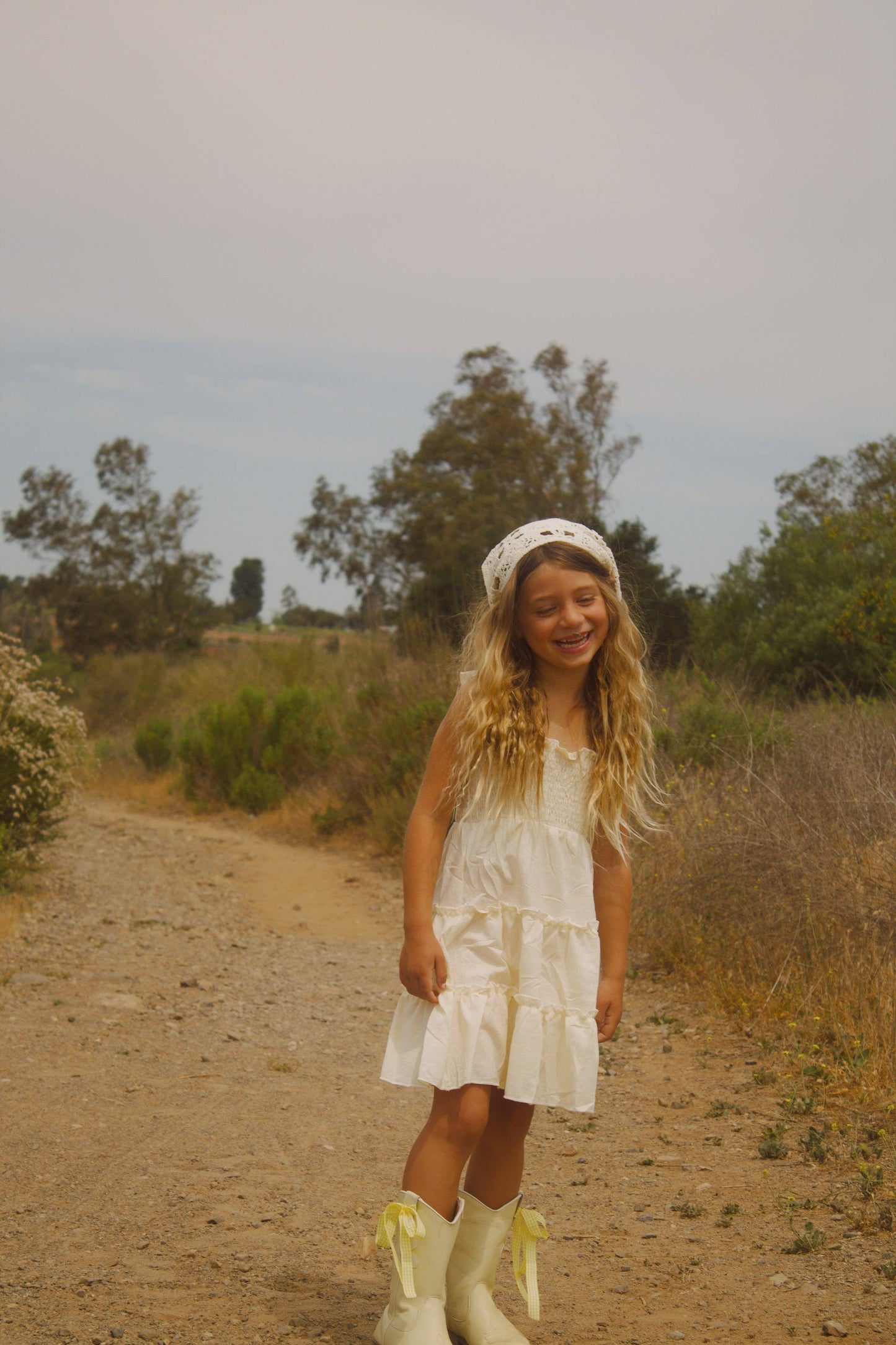 Young girl in a white dress and rain boots standing on a dirt path with trees in the background
