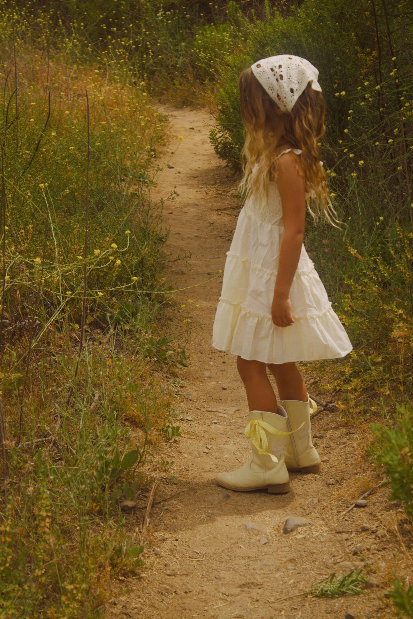 Young girl in a white dress and bonnet walking along a dirt path in a natural setting.