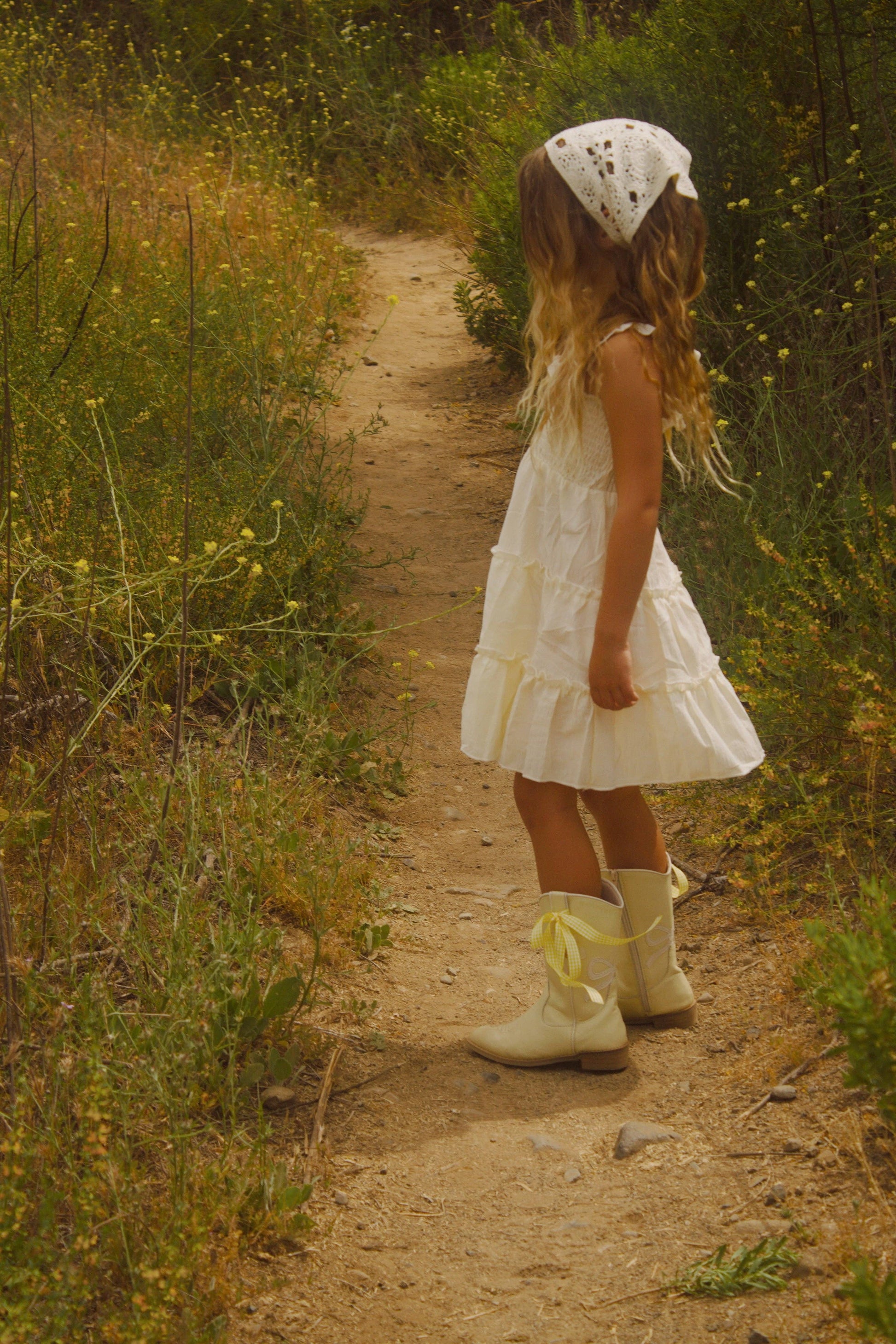 Young girl in a white dress and bonnet walking along a dirt path in a natural setting.