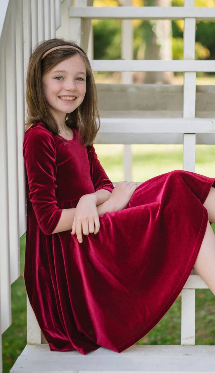 Young girl in a red velvet dress sitting on a white bench outdoors.