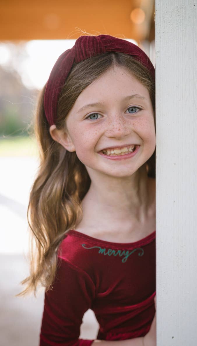Young girl wearing a red headband and dress with 'mary' on it, standing against a white wall.
