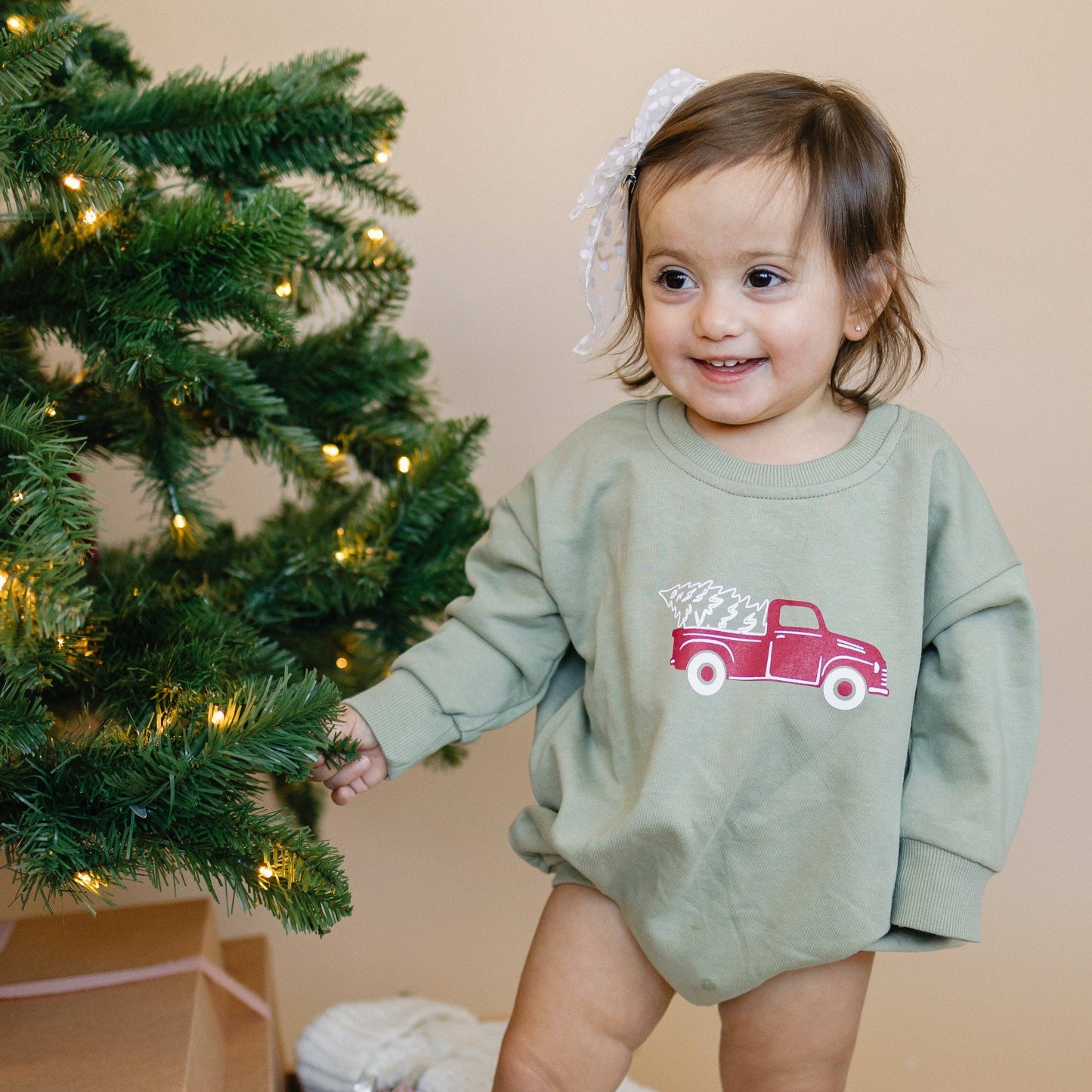 Child wearing a green sweatshirt with a red truck design, standing next to a decorated Christmas tree.