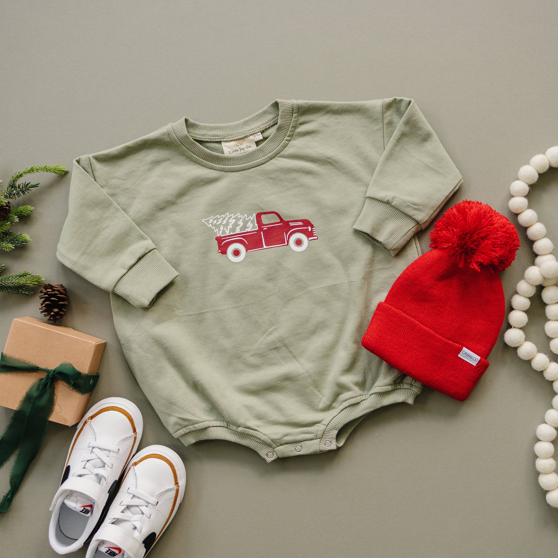 Green baby onesie with red truck design, red beanie, white sneakers, and small gift on a gray surface.
