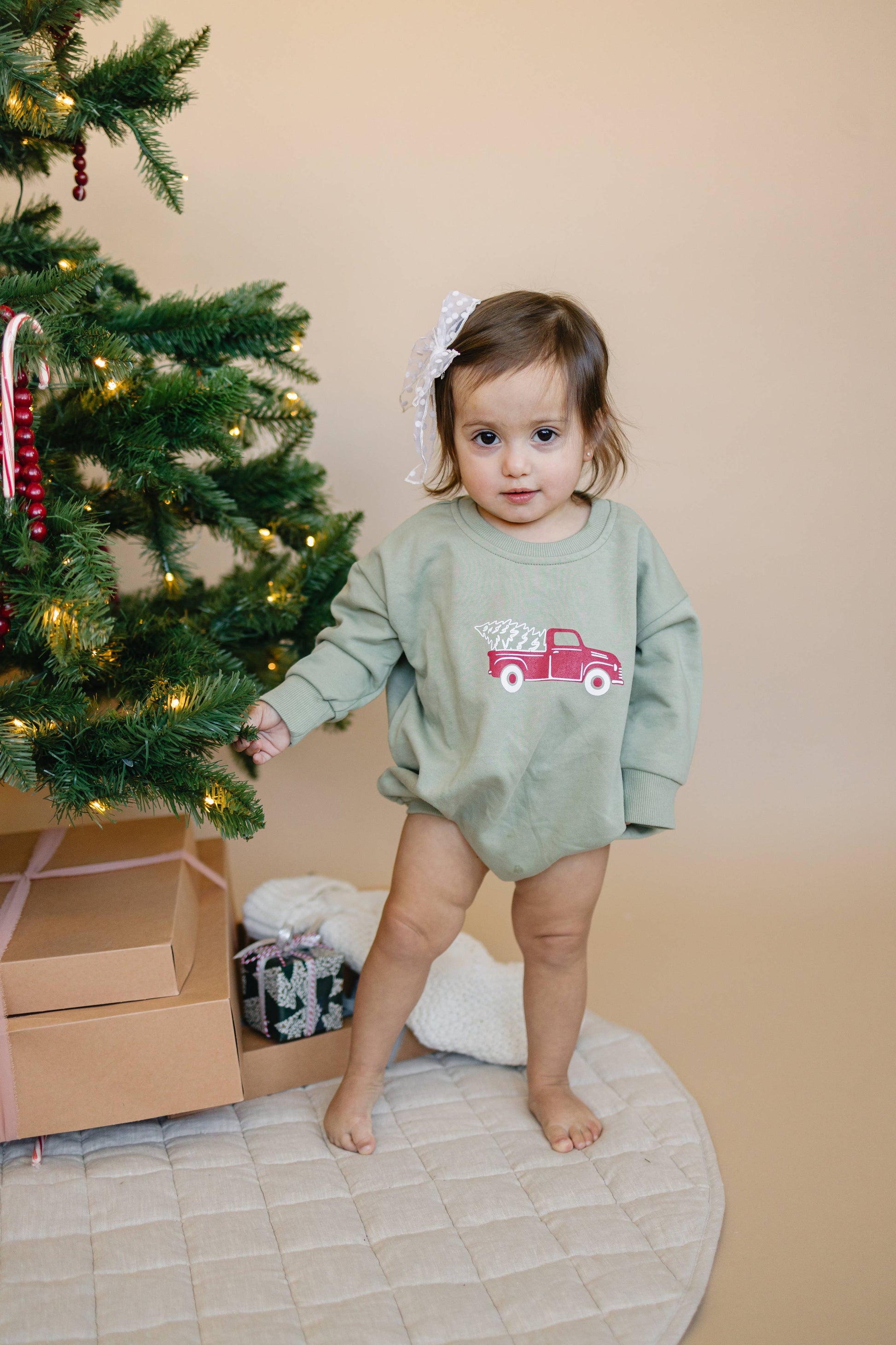 Child in a green onesie with a red truck design standing next to a decorated Christmas tree.