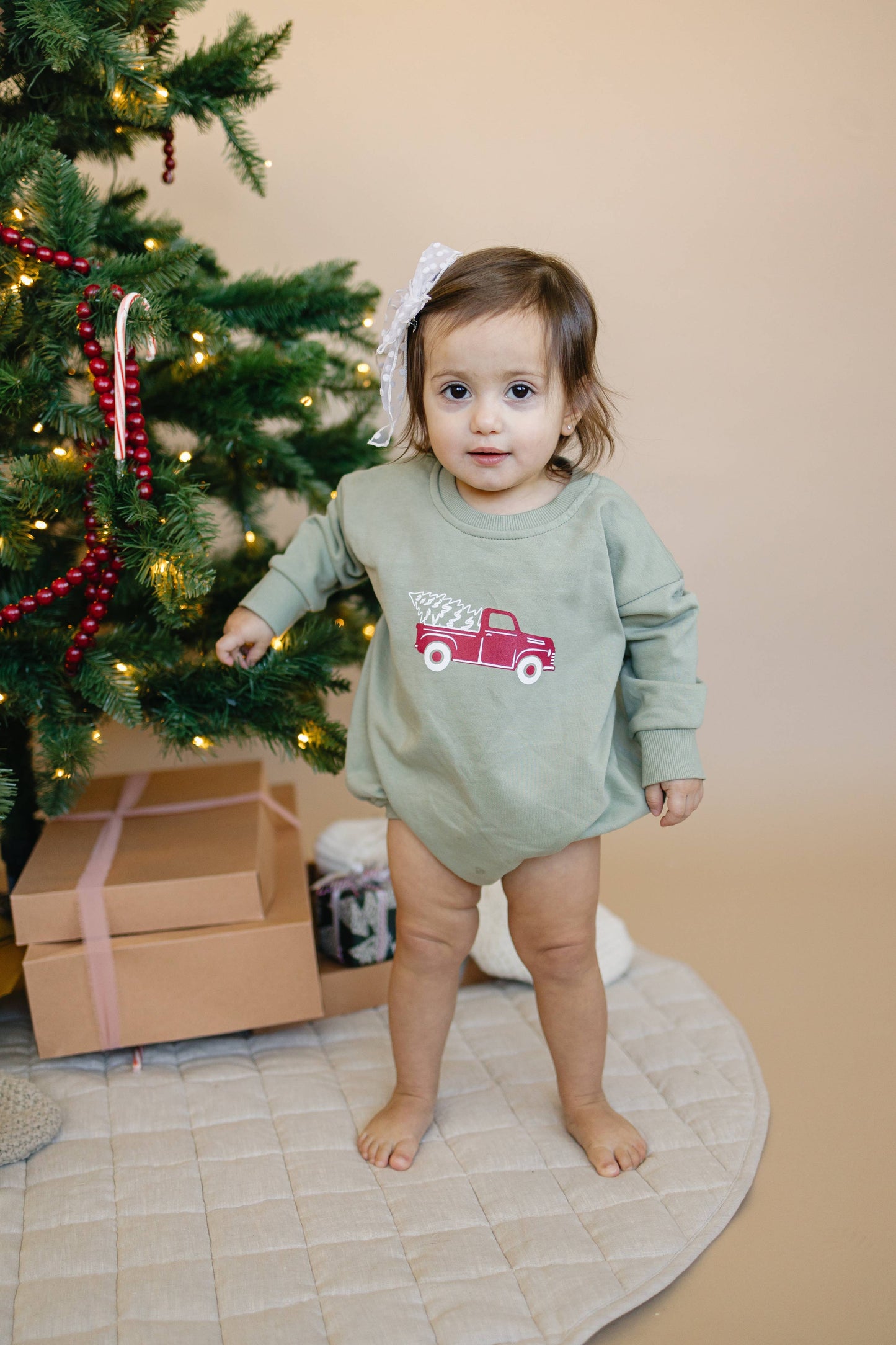 Child in a green outfit with a red truck design standing next to a decorated Christmas tree.