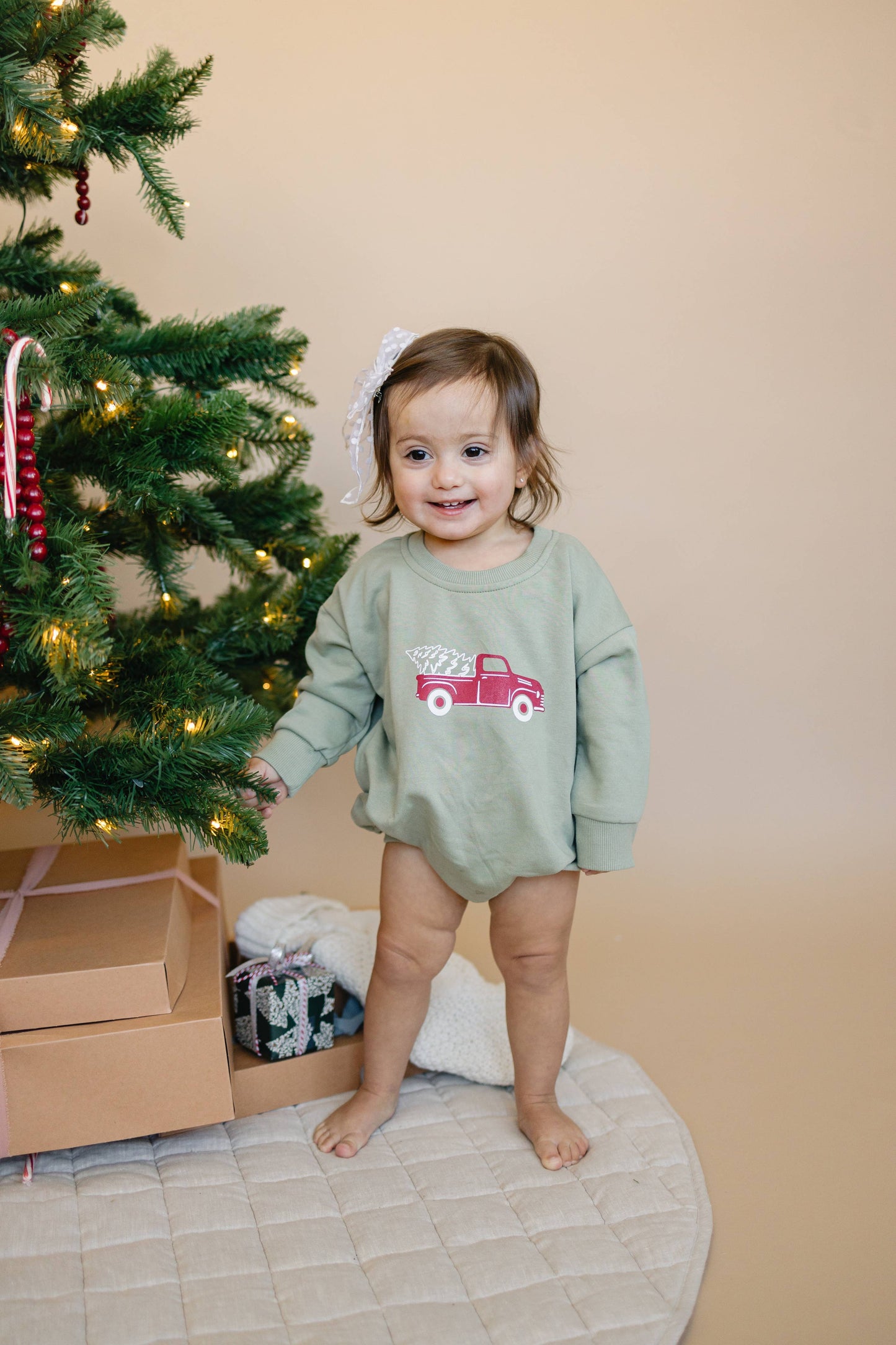 Child in a green outfit with a red truck design standing next to a decorated Christmas tree.