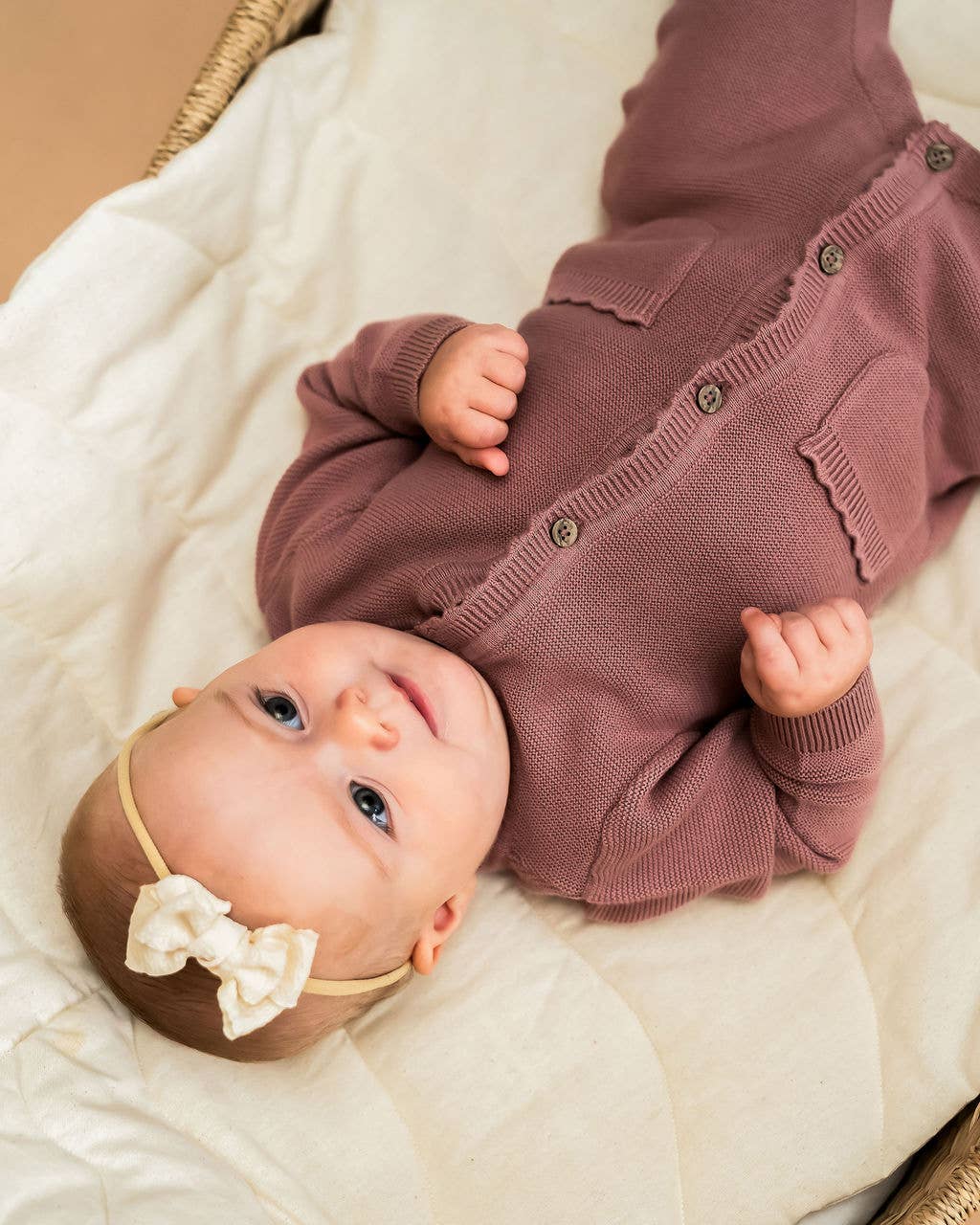 Baby wearing a mauve outfit with a bow headband on a white blanket