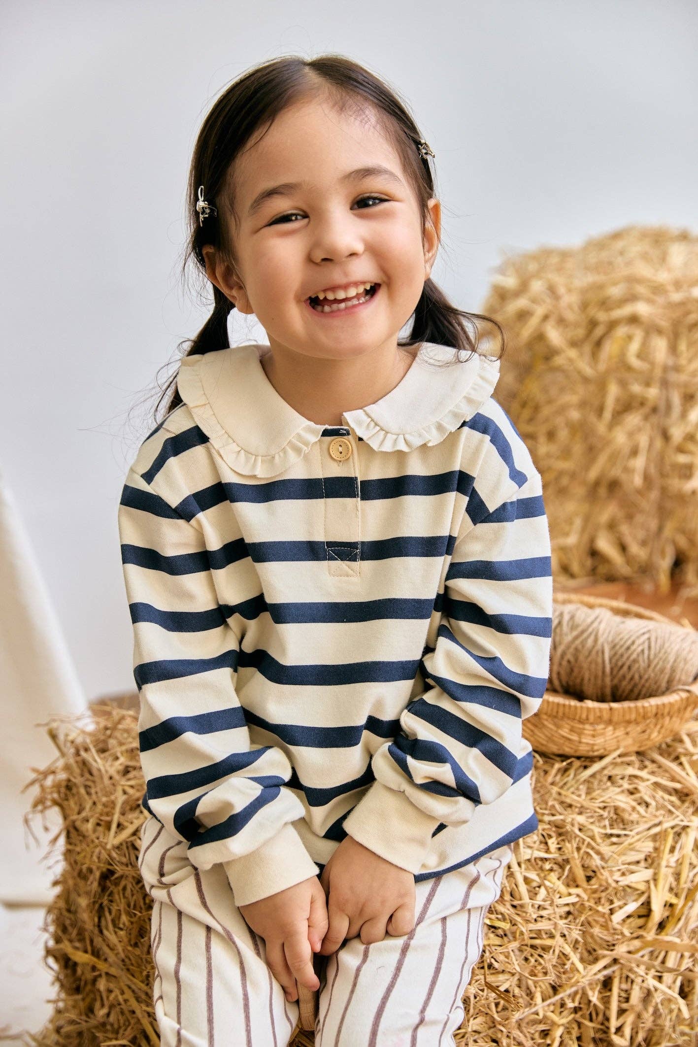 Child wearing a striped shirt with a white collar, standing on hay bales.