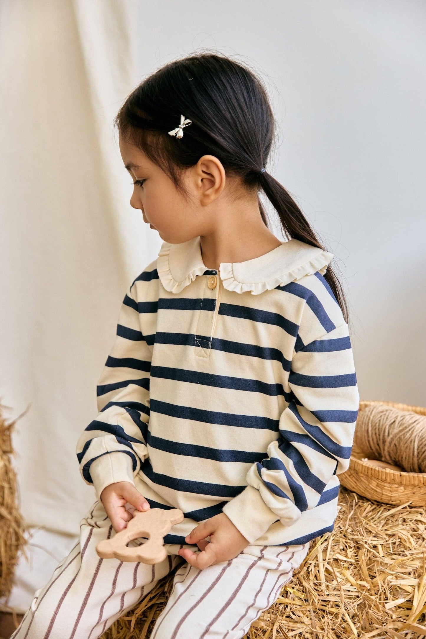 Young girl wearing a striped shirt with a white collar, sitting on straw with a neutral background