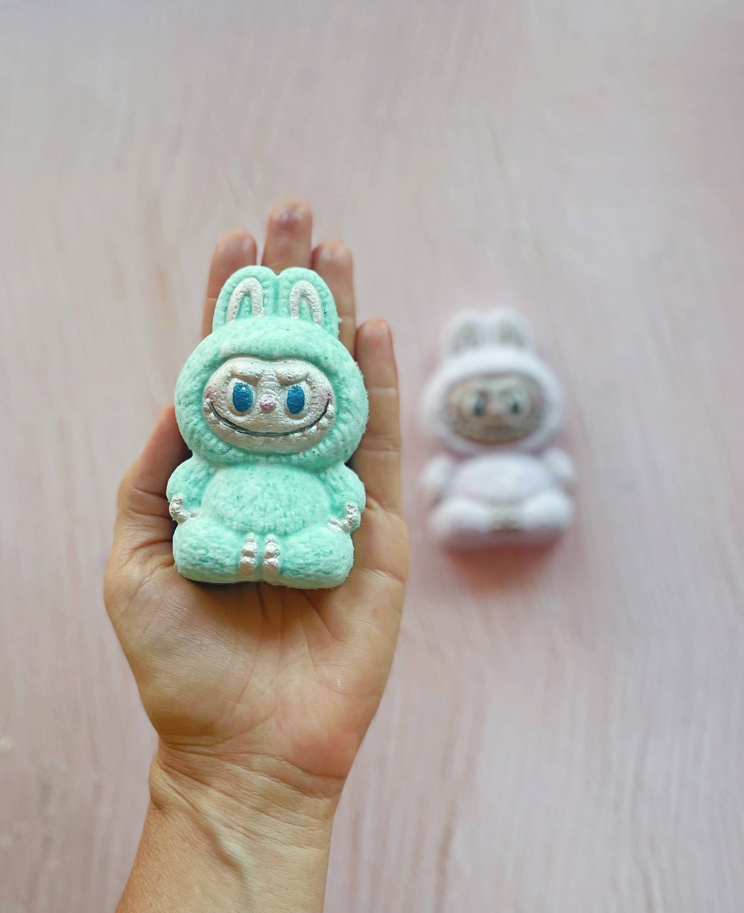 Small green plush toy with bunny ears held in a hand on a light wooden surface.