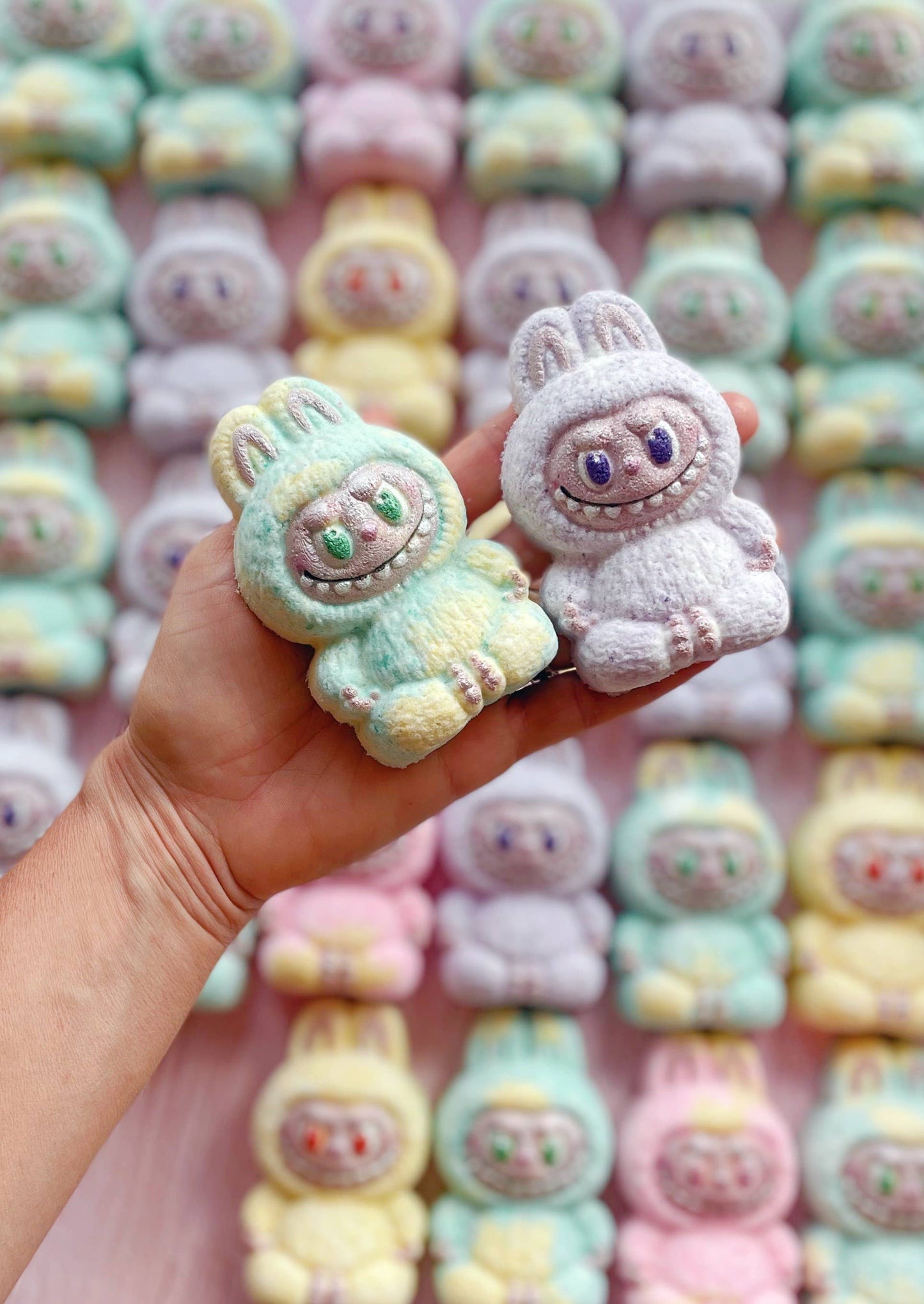 Pastel-colored plush toys resembling cute characters held in a hand with a pink background.