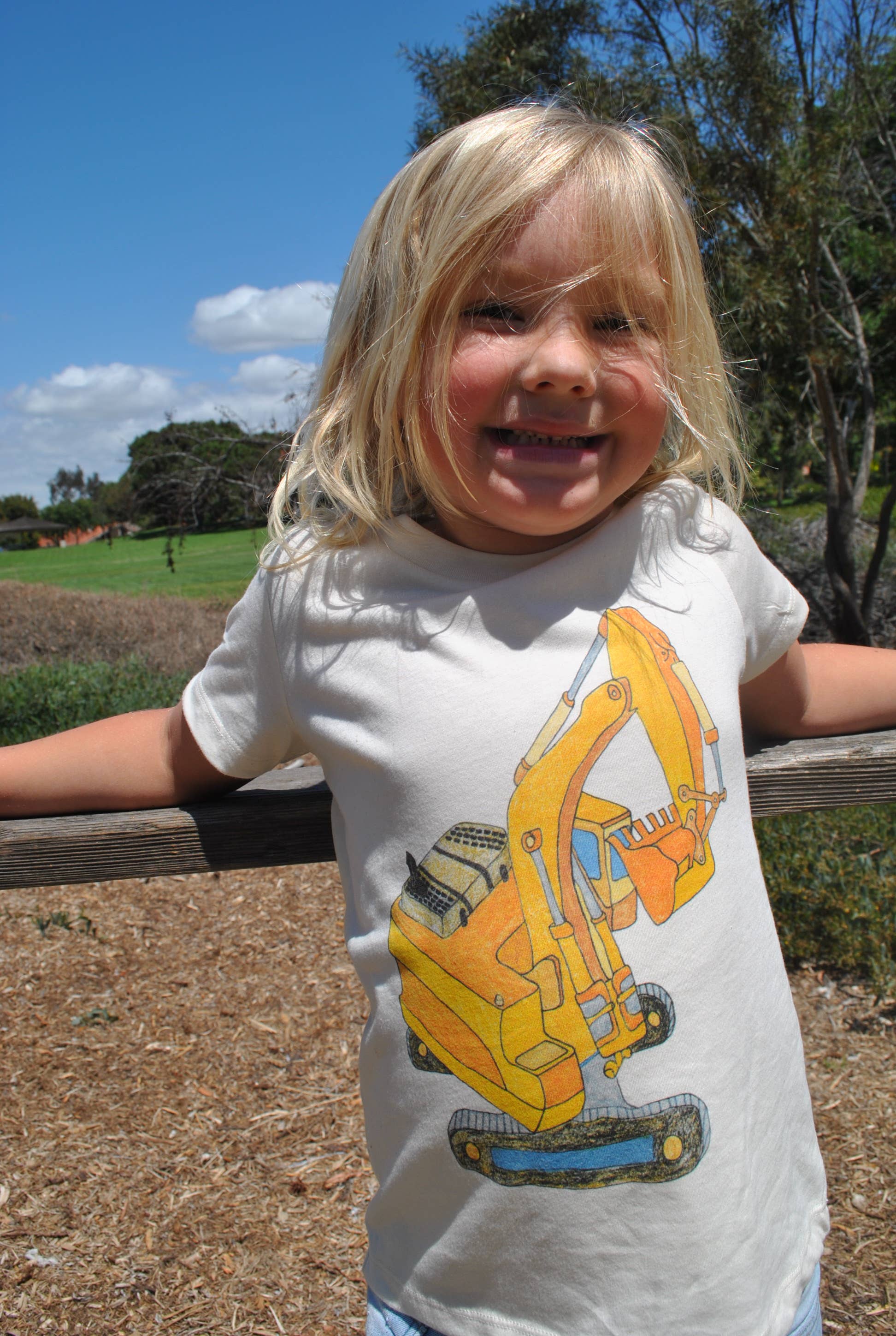 Child wearing a white t-shirt with a yellow excavator design, standing outdoors.