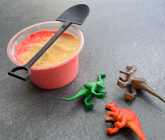 Container of red and yellow substance with a black spoon, surrounded by toy dinosaurs on a gray surface.