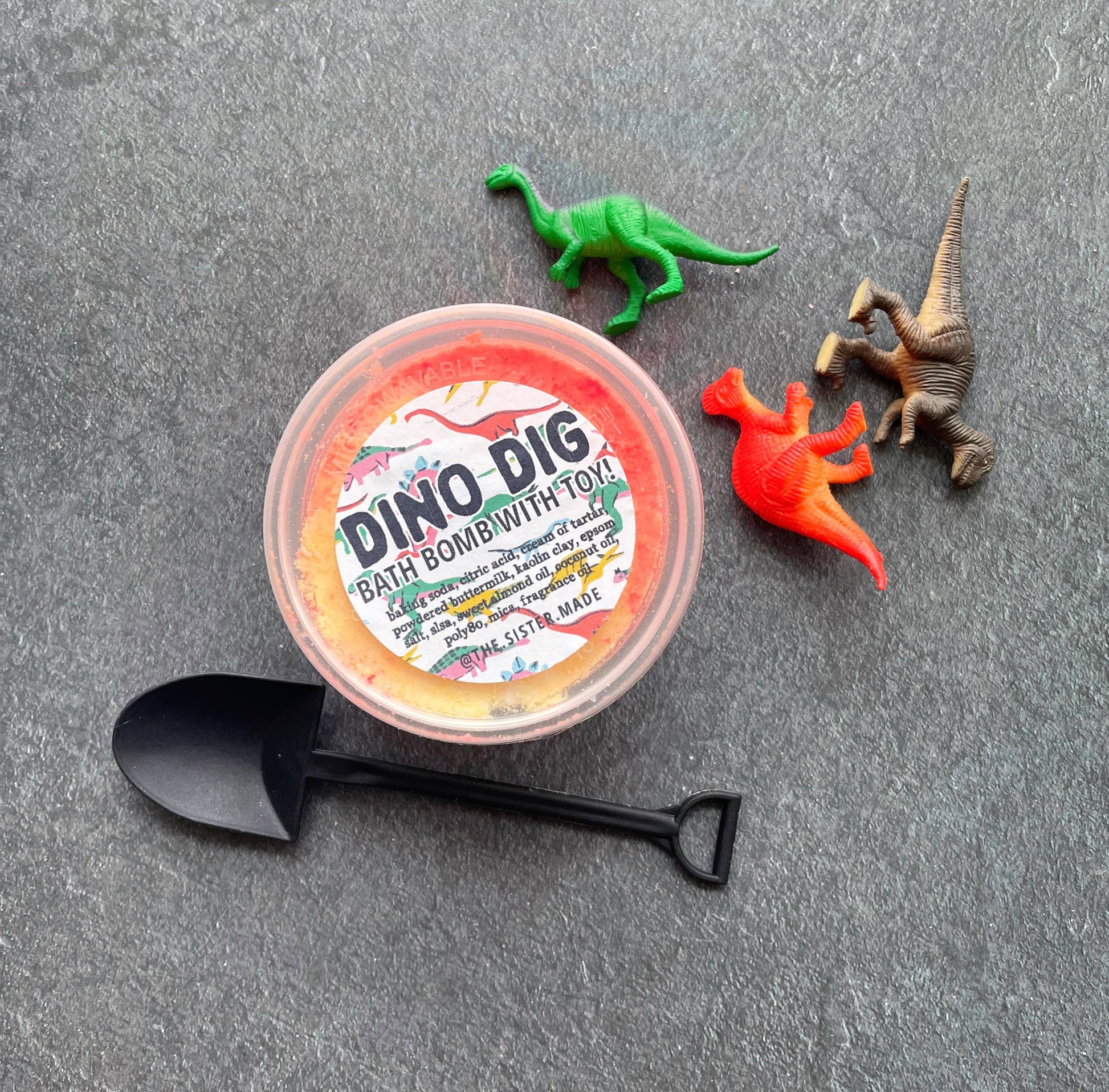 Dino Dig bath bomb with toy on a gray surface with toy dinosaurs and a black scoop.