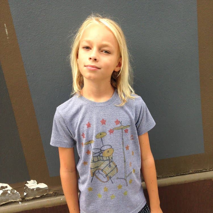 Young girl wearing a blue t-shirt with a graphic design against a gray wall.