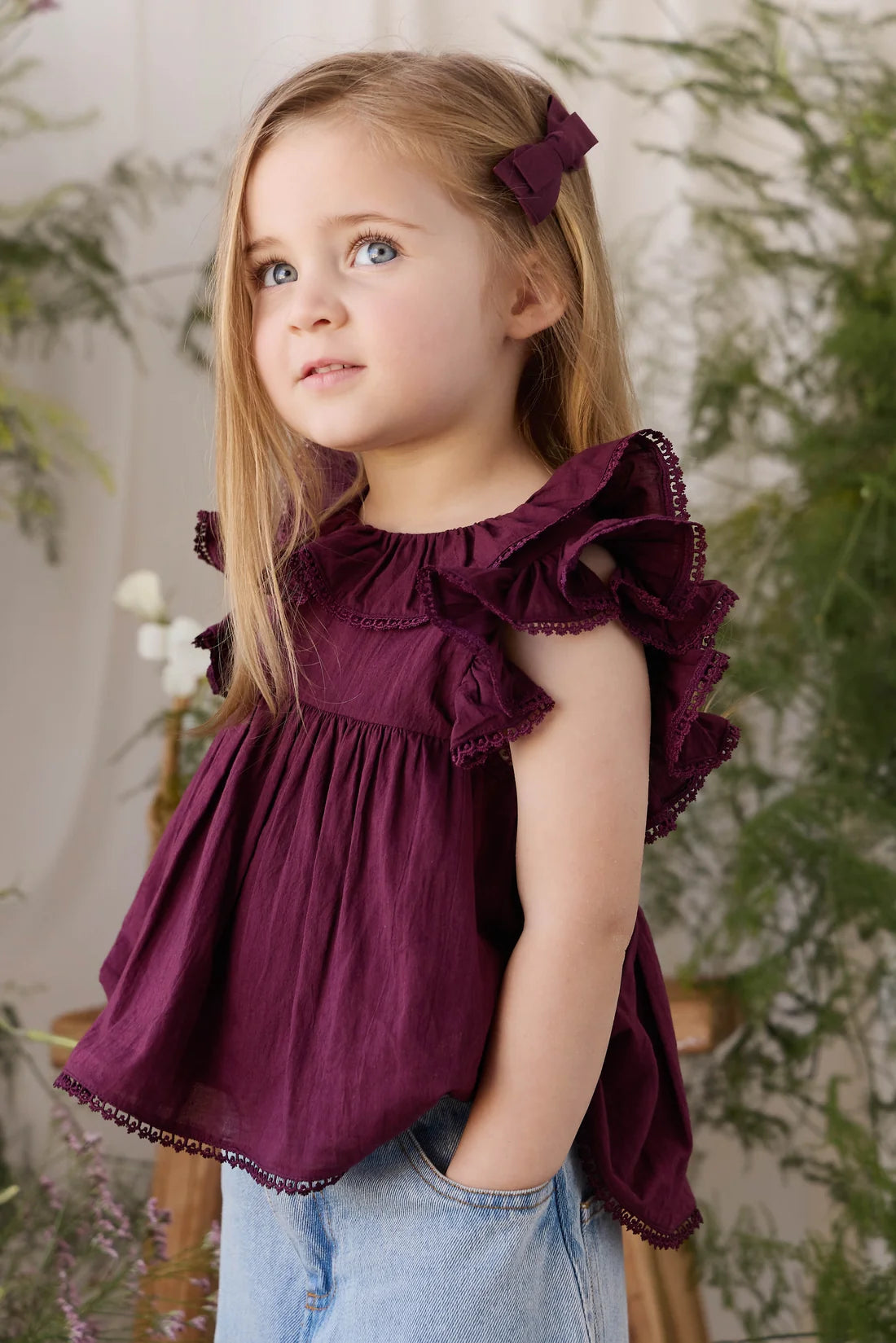 A young girl with blonde hair wearing a deep burgundy ruffled sleeveless top and light blue jeans, standing in front of a soft-focus indoor background with green plants.