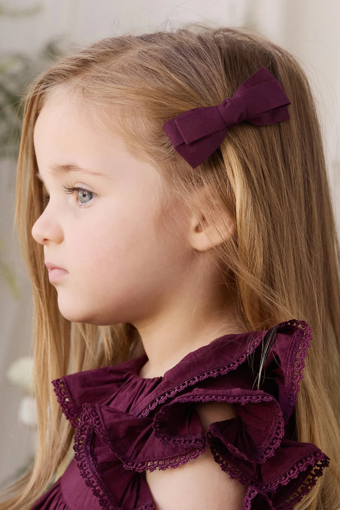 A young girl with long blonde hair wearing a deep purple bow hair clip and a matching purple ruffled top, shown in profile against a soft light background.