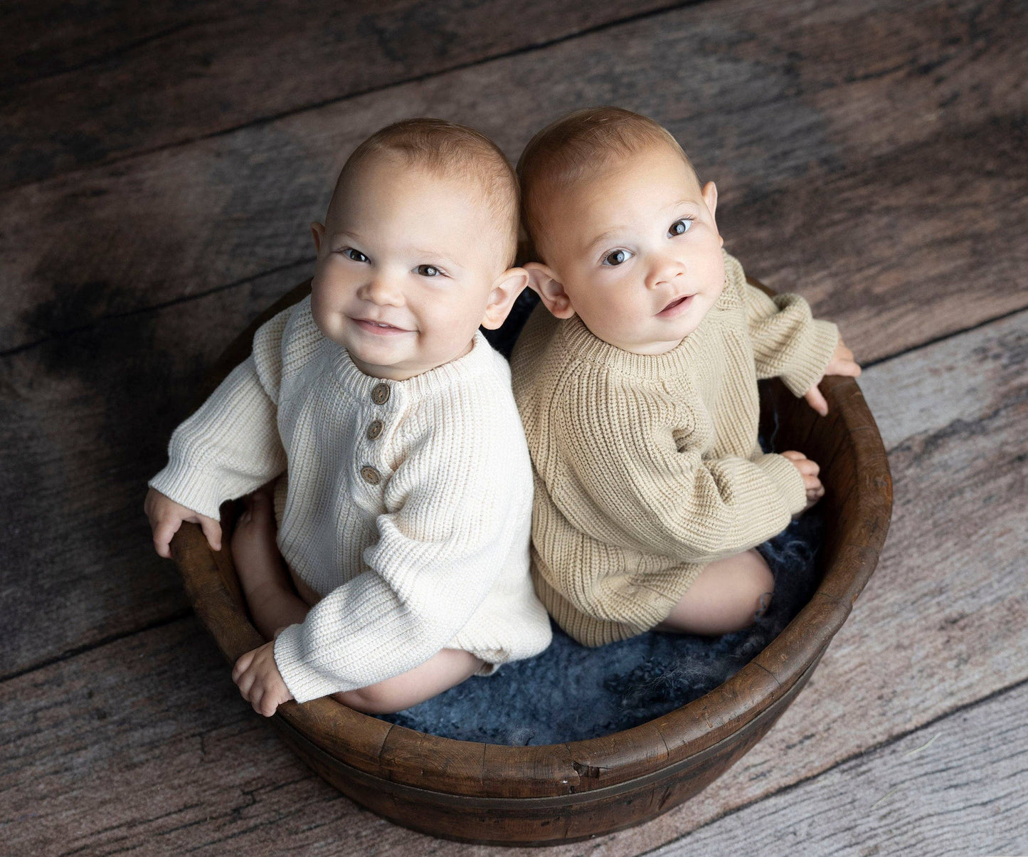 Two babies in knitted outfits sitting in a wooden basin on a rustic wooden floor.