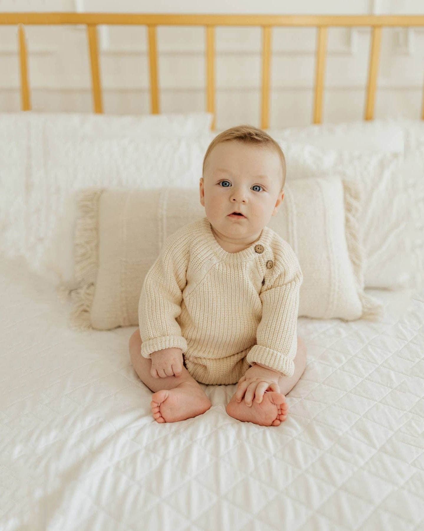 Baby sitting on a bed wearing a beige ribbed outfit.