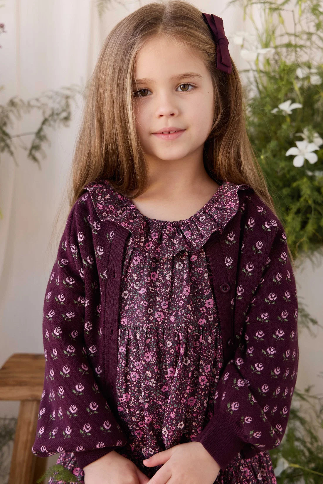 A young girl with long light brown hair wearing a dark purple floral dress with a matching hair bow, standing in front of a softly blurred indoor background with greenery.