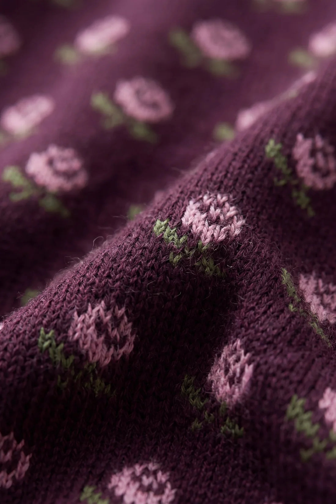 Close-up of a knitted fabric in deep plum purple with a repeating pattern of pink flowers and green stems.