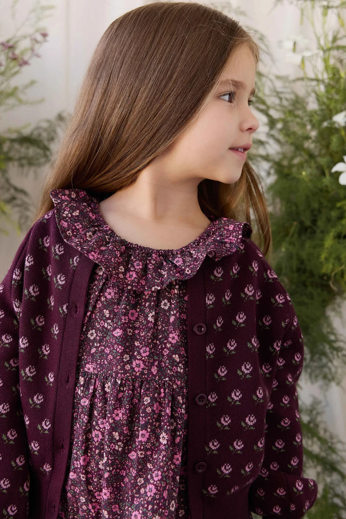 A young girl with long brown hair is shown in profile wearing a dark purple floral cardigan over a matching floral top. The background features soft-focus green foliage and a light wall.