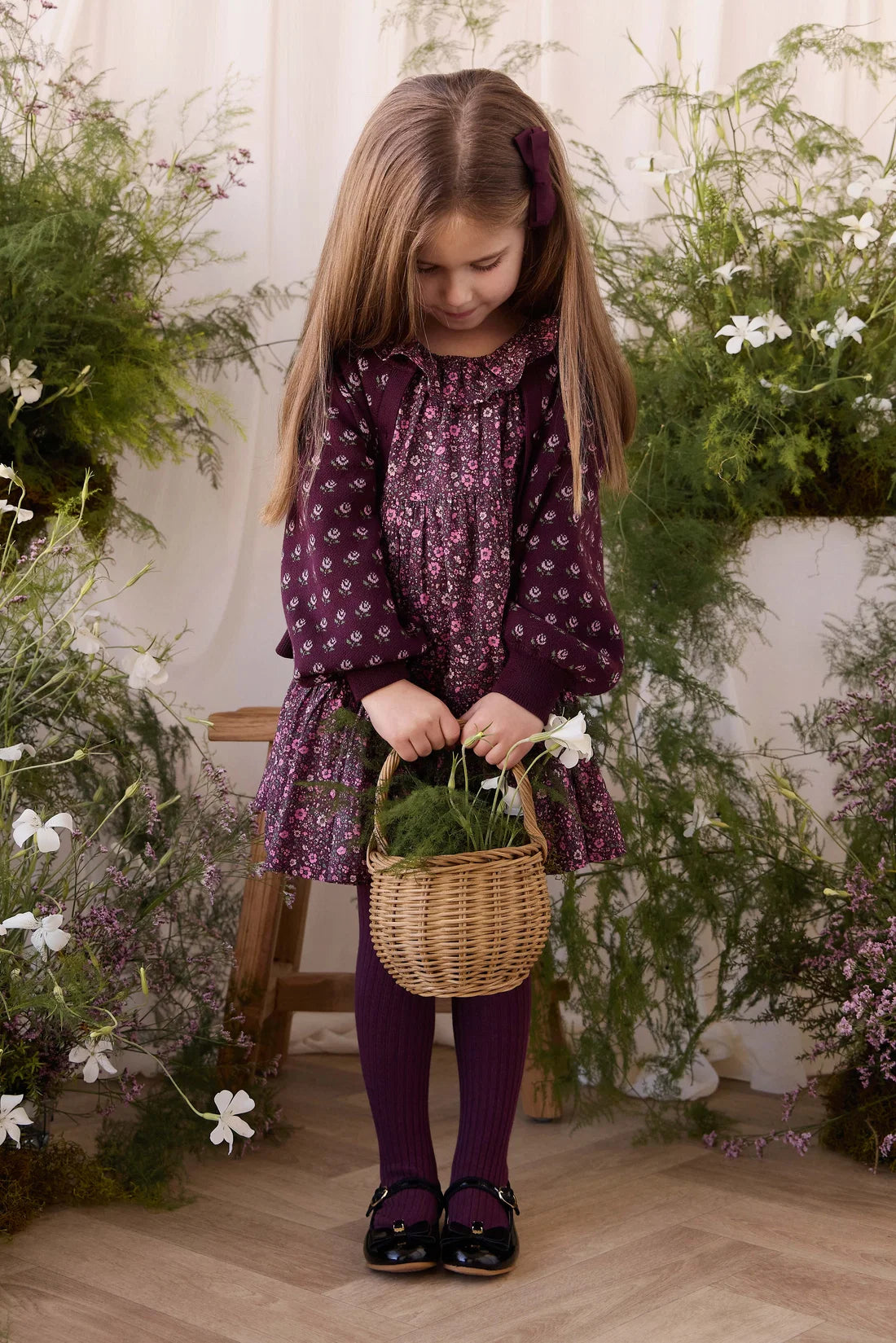 A young girl with long light brown hair stands indoors holding a small wicker basket with green stems. She is wearing a dark purple patterned long-sleeve dress, matching purple tights, and black shoes.