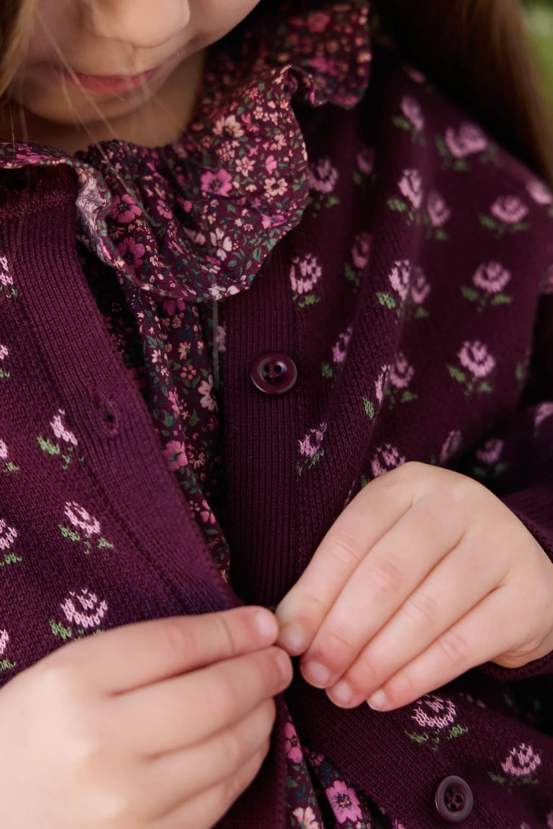 Close-up of a child wearing a dark purple knit cardigan with a pink and green floral pattern, fastening a button with both hands.