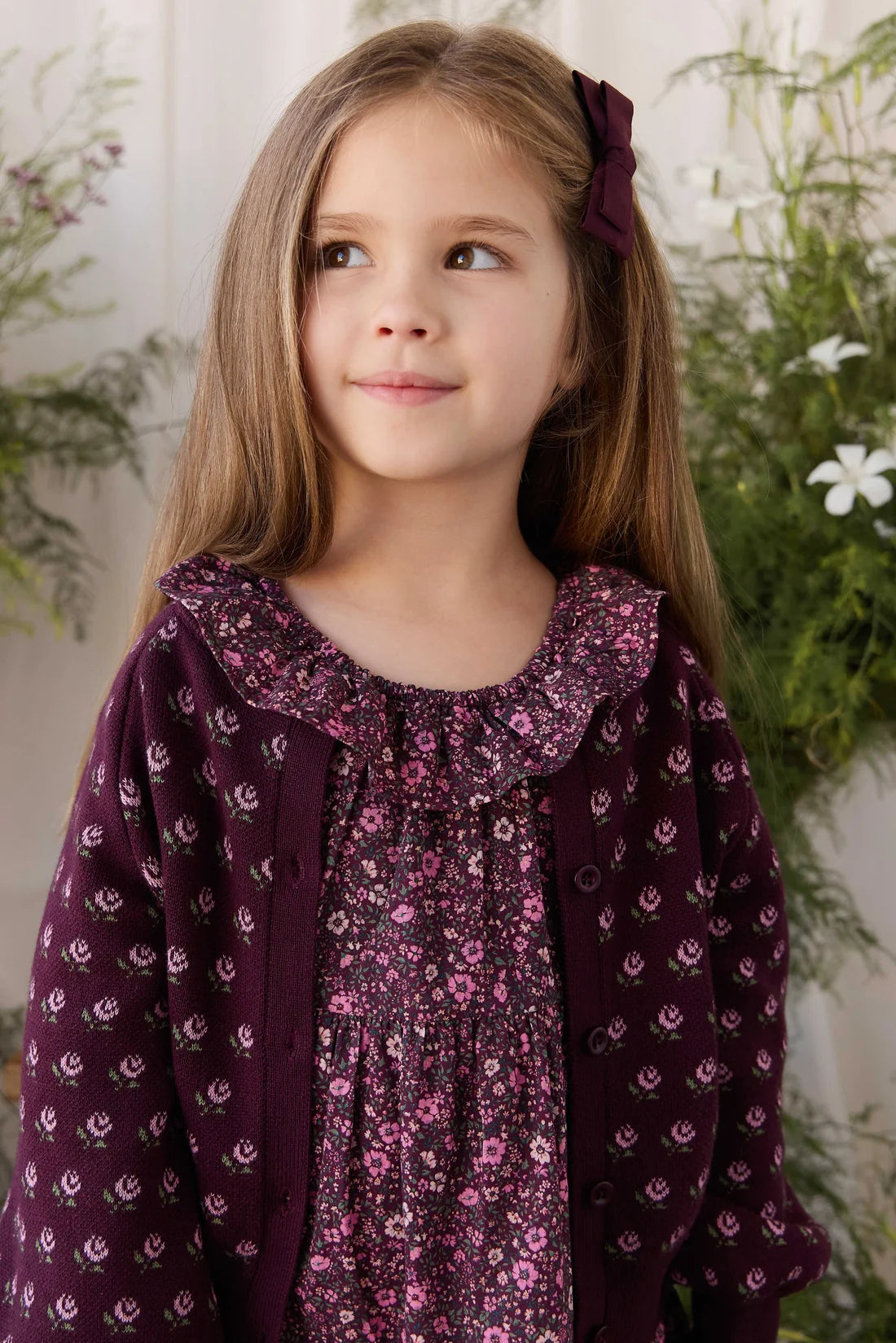 A young girl with long light brown hair wearing a dark purple floral dress and matching patterned cardigan, with a matching hair bow, set against a soft-focus background of green foliage and white flowers.