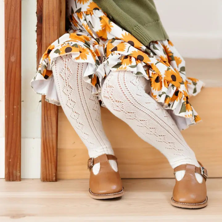 Person wearing a floral skirt, white lace pants, and brown shoes sitting on a wooden floor.