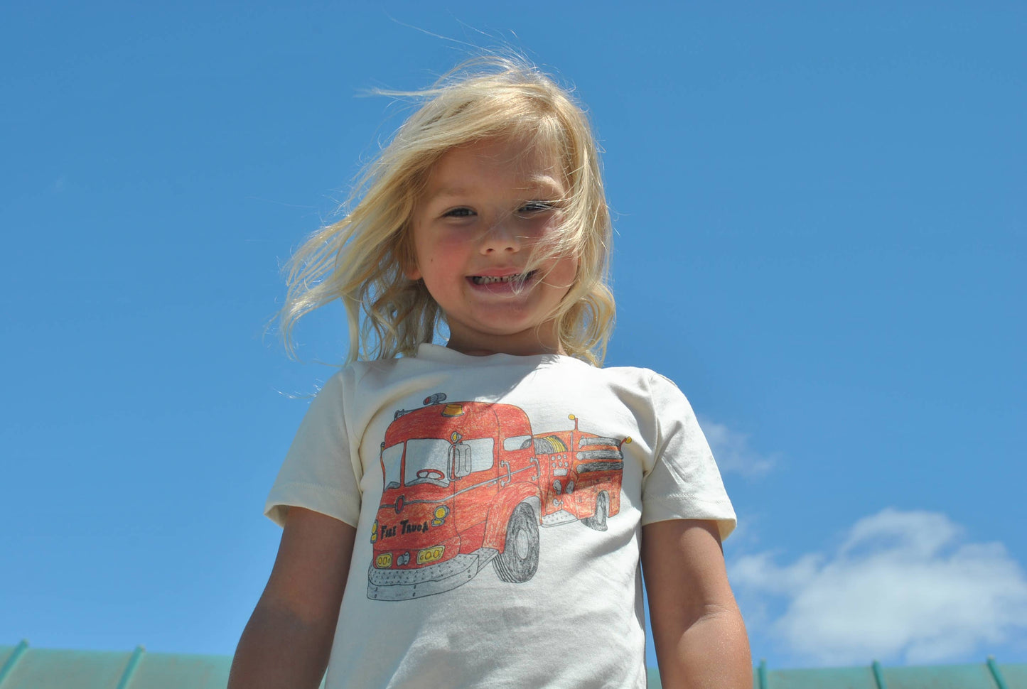 Child wearing a t-shirt with a fire truck design against a blue sky