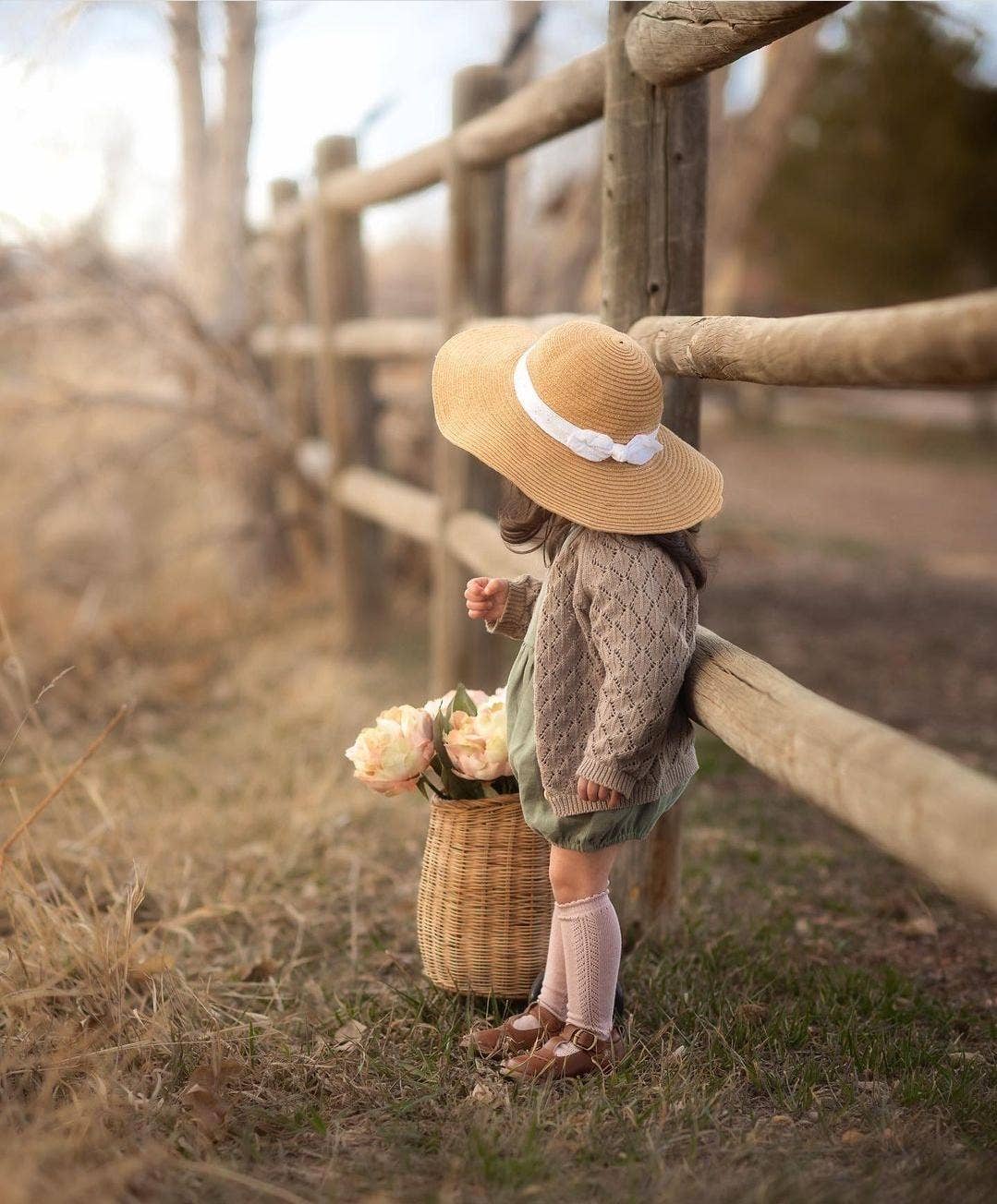 Child in a straw hat with a basket of flowers standing near a wooden fence.