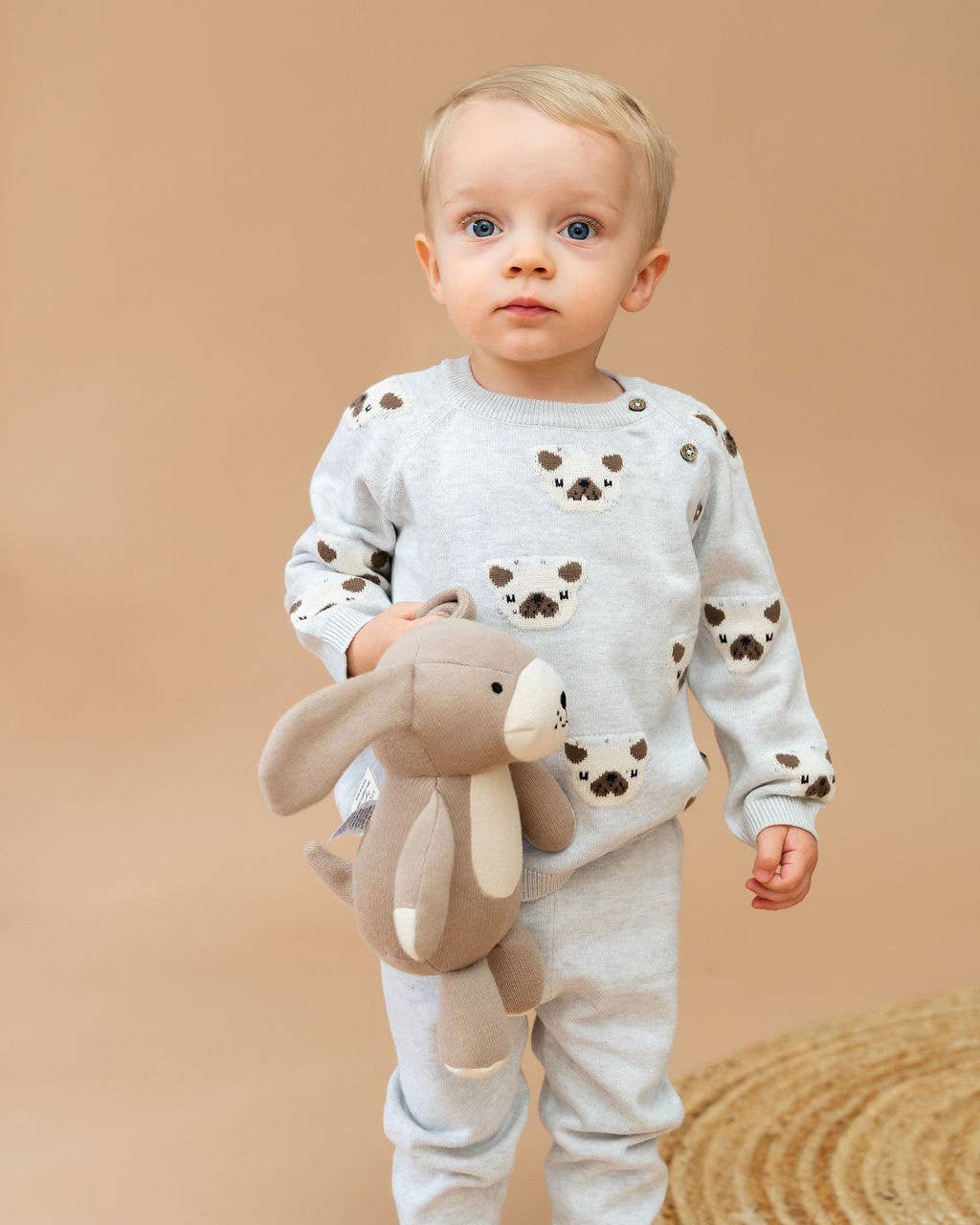 Baby wearing a white outfit with brown animal prints, holding a plush toy against a beige background