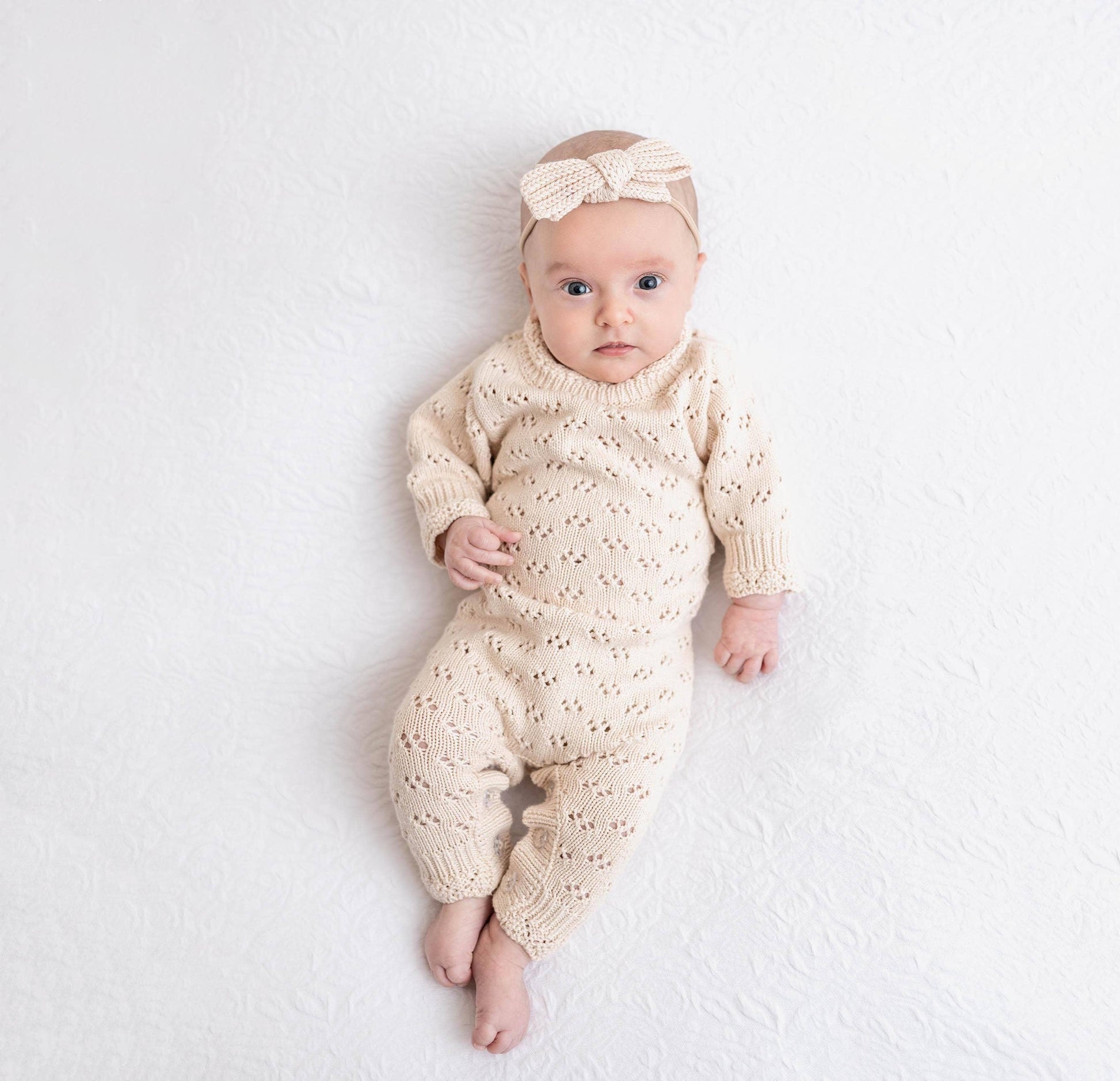 Baby wearing a cream knitted outfit and headband on a white background