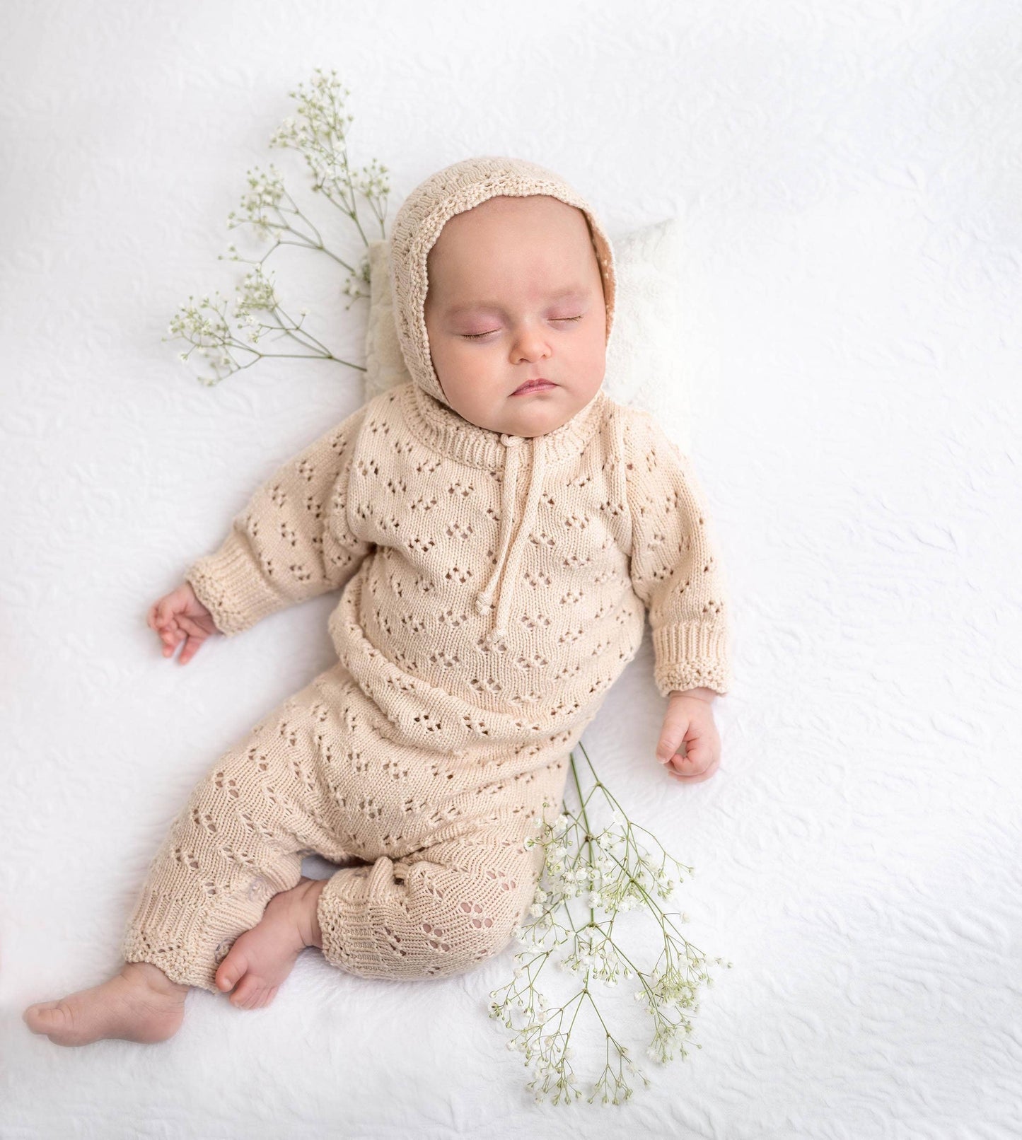 Baby in a beige knitted outfit lying on a white surface with small green plants.