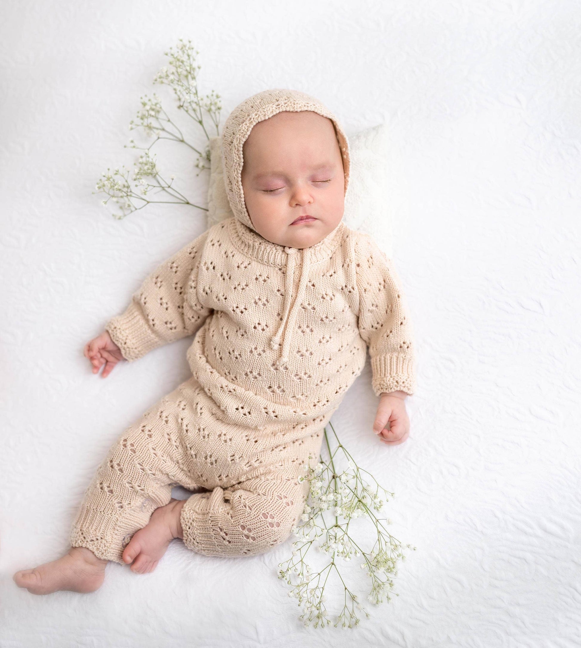 Baby in a beige knitted outfit lying on a white surface with small green plants.