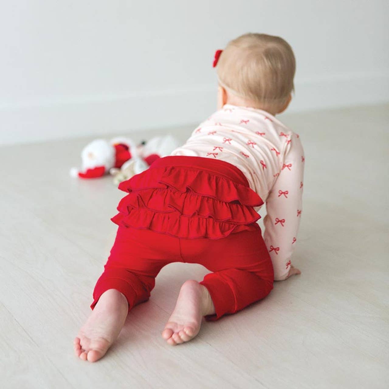 Baby wearing a pink outfit with red ruffled pants sitting on a light-colored floor.