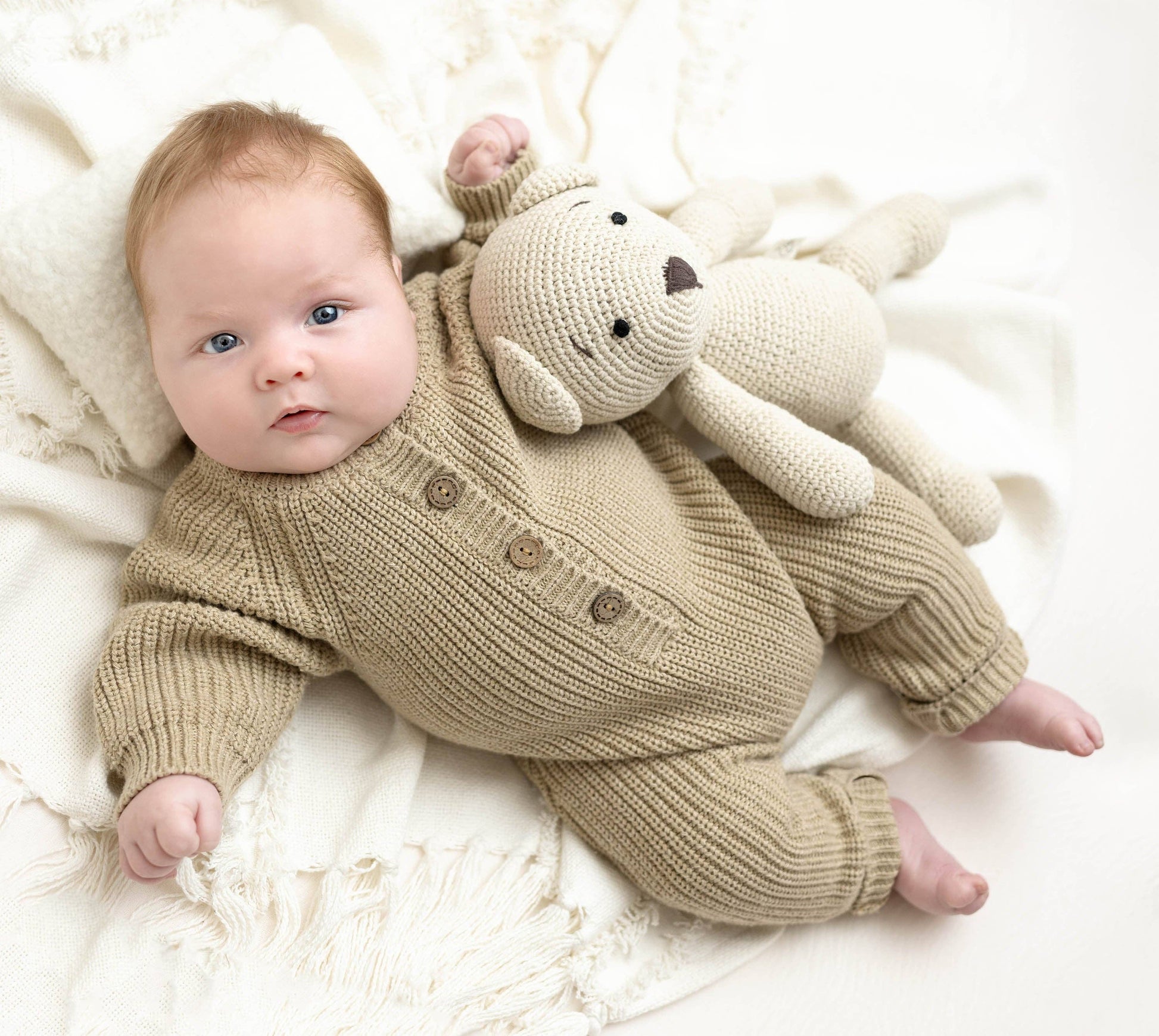 Baby in a beige knitted outfit lying on a white blanket with a plush toy.